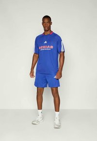 Vyras stovi dėvėdamas mėlyną Adidas sportinį marškinėlį ir šortus, baltas kojines ir baltus sportinius batus prieš paprastą foną.