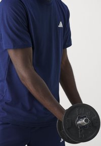 Marinblå kortärmad sporttröja med vitt logotyp, hålls av en muskulös arm som greppar en svart justerbar hantel.