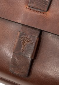 Brązowa skórzana torba z teksturowaną powierzchnią, wyraźnym przeszyciem i wytłoczonym dekoracyjnym zapięciem z motywem roślinnym.