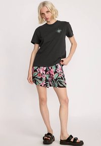 Czarny bawełniany t-shirt z nadrukiem turkusowego kompasu, w połączeniu z czarnymi szortami ozdobionymi żywym różowo-zielonym wzorem kwiatowym. Czarne sandały.