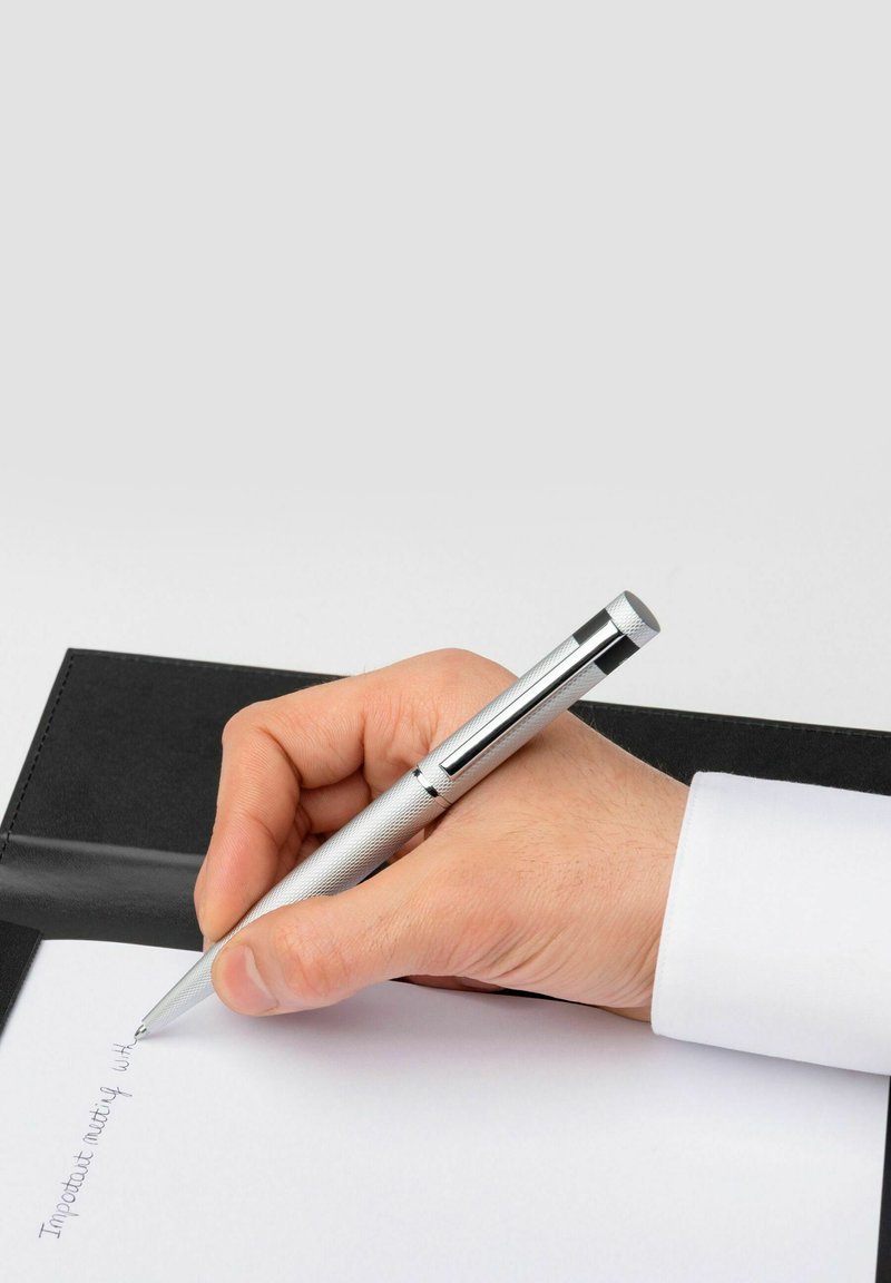Penna in metallo argento con impugnatura testurizzata tenuta in una mano, mentre scrive su carta bianca posta su una copertina nera in pelle di un taccuino.