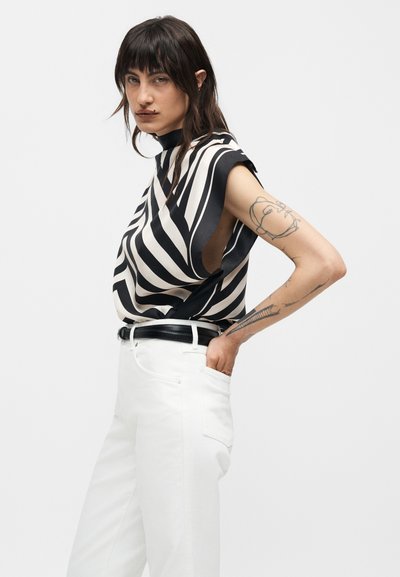 Γυναίκα με σκούρα μαλλιά που φοράει ασπρόμαυρο ριγέ αμάνικο τοπ και λευκό παντελόνι, δείχνοντας τατουάζ στο δεξί της χέρι.