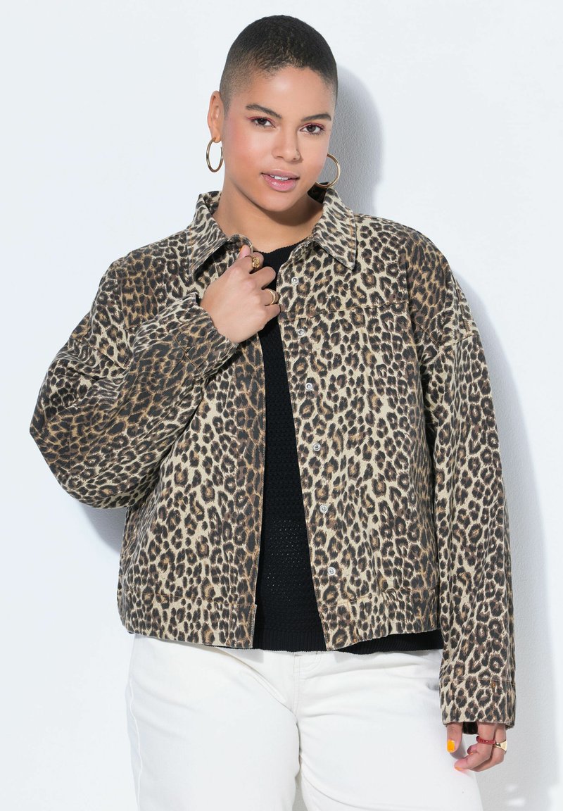 Person med kort hår iført store øreringe i hoop-stil, en leopardprint jakke med knapper over en sort top og hvide bukser, poserer mod en hvid baggrund.