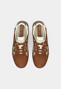 Καφέ αθλητικά παπούτσια από σουέτ με κρεμ λεπτομέρειες, διαθέτοντας στρογγυλεμένη μύτη, δέσιμο με κορδόνια και λογότυπο στη γλώσσα. Υφή στην επιφάνεια και αντίθετες ρίγες.