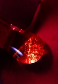 Blaues Flammenrohr erwärmt einen kleinen Metalltiegel mit einer leuchtend schmelzenden Substanz darin, vor einem dunkelroten Hintergrund.