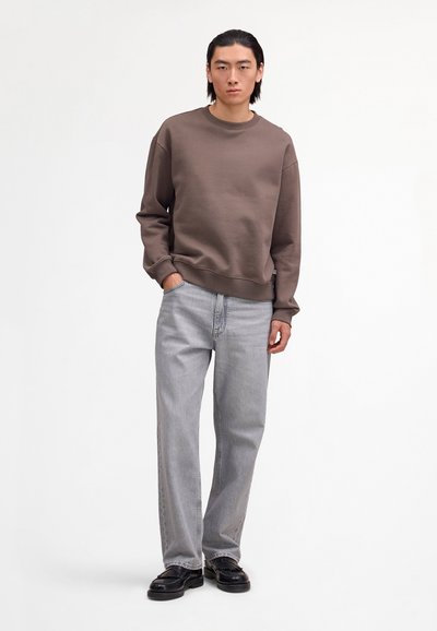 Homme portant un sweat-shirt marron, un jean gris clair et des mocassins noirs, debout avec une main dans la poche contre un fond blanc uni.