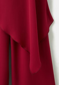 Czerwona tkanina z warstwowym, asymetrycznym dołem i gładką fakturą, zawieszona na białym tle.