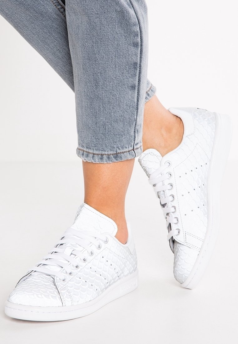 Zapatillas blancas hechas de un material texturizado con un patrón de escamas, con cordones blancos y ojales de metal, combinadas con jeans cortos azules.