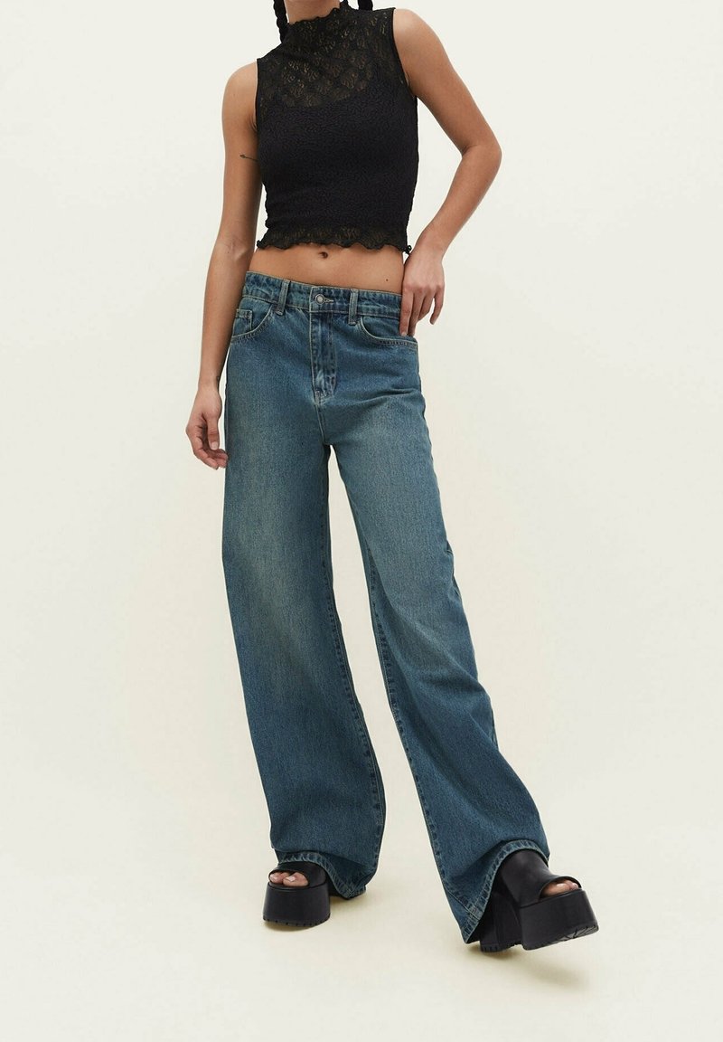 Top corto de encaje negro con bordes festoneados, combinado con jeans de mezclilla azul de pierna ancha y sandalias de plataforma negras y gruesas. Diseño simple, tejidos texturizados.