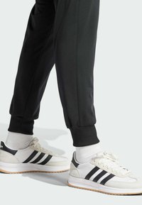 Weiße Sportlichkeitsschuhe mit schwarzen Streifen und Gummisohlen, kombiniert mit schwarzen Jogginghosen und weißen Crew-Socken. Die Schuhe haben Suede-Akzente.