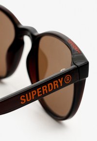 Solbriller med en stel i skildpadde-tekstur, brune linser og en orange "SUPERDRY"-logo på siden. Vinklet design med polerede detaljer.