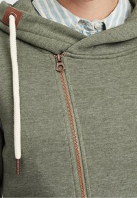 Sudadera verde con cremallera y acabado texturizado, que presenta una cremallera de color cobre en contraste y cordones crema. Camisa a rayas azul visible debajo.
