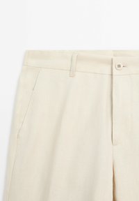 Pantalon en lin beige avec devant plat, poches latérales et fermeture à un bouton. Texture lisse avec une finition en tissu léger.