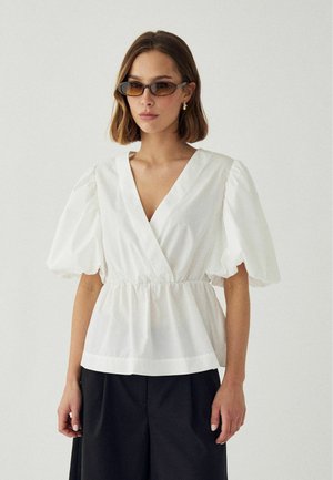 Žena s jantárovo sfarbenými slnečnými okuliarmi, biela blúzka s nafúknutými rukávmi a výstrihom do V, čierne široké nohavice, stojaca pred jednoduchým pozadím.