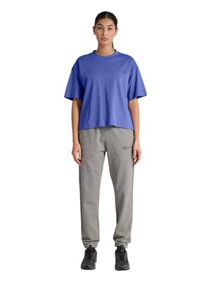 Kvinde stående med fronten fremad iført en løs blå kortærmet T-shirt, grå joggingbukser og sorte sneakers på en hvid baggrund.