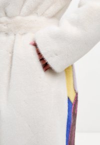 Faux fur-kappa i vitt med mjuk textur. Har en ficka, breda ärmar och färgglada geometriska detaljer i gult, blått, rosa och brunt.