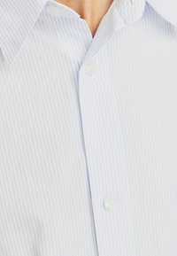 Ανοιχτό μπλε πουκάμισο με κουμπιά φτιαγμένο από ύφασμα με λεπτές κάθετες άσπρες ρίγες, διαθέτοντας κλασικό γιακά και κουμπιά που μοιάζουν με πέρλες.