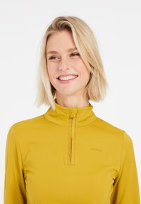 Dlouhý rukáv, zipová blůza v hořčicově žluté barvě, s vysokým límcem a malým logem. Materiál má hladkou, elastickou strukturu.