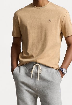 Camiseta de algodón color beige con cuello redondo clásico y un pequeño logotipo en el pecho; combinada con pantalones de chándal gris claro con cordón en la cintura.