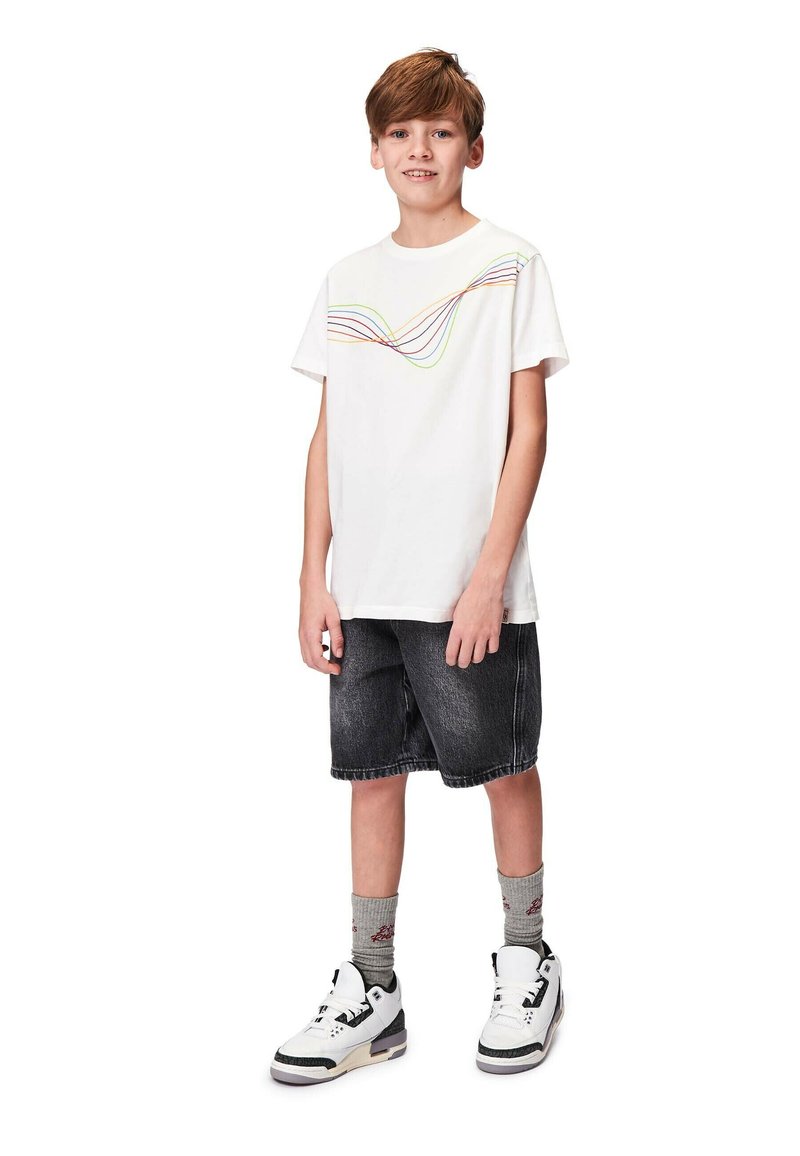 Vit bomulls-t-shirt med ett färgglatt vågmönster över bröstet, kombinerad med svarta denimshorts och vita sneakers med grå strumpor.