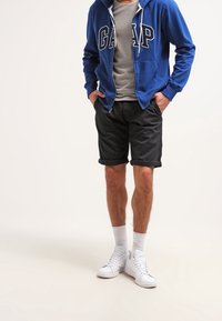 Persona che indossa una felpa blu GAP, una maglietta grigia, pantaloni corti neri arrotolati, calzini bianchi e sneaker bianche, in piedi con le mani nelle tasche.