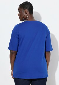Blauwe T-shirt met korte mouwen, gemaakt van een gladde stof. Heeft een ronde halslijn en een ontspannen pasvorm, gecombineerd met donkere broek en grote hoopsieraden.