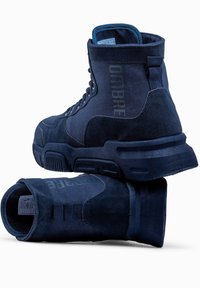 Bottes montantes bleu marine en suede et en tissu, avec une semelle en caoutchouc robuste, un laçage à l'avant et un détail de marque sur les côtés.