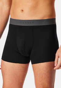 Schwarze Boxershorts aus weichem Stoff mit einem grauen elastischen Bund, auf dem der Markenname "SCHIESSER" in Grau eingeprägt ist.