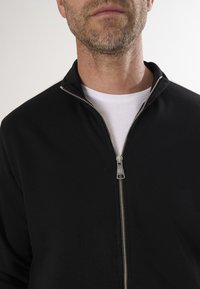 Schwarze Zip-Jacke aus einem weichen Stoff, mit Stehkragen und silbernen Reißverschlussdetails, getragen über einem weißen T-Shirt.