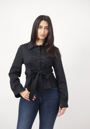 Femme aux longs cheveux noirs portant une chemise noire boutonnée nouée à la taille et un jean bleu foncé, debout devant un fond blanc uni.