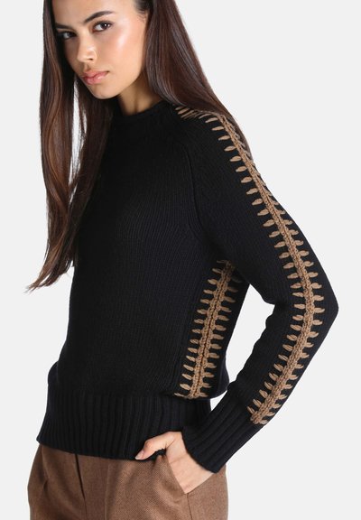 Femme aux longs cheveux bruns portant un pull noir avec un motif tricoté marron sur les épaules et les manches, les mains dans les poches d’un pantalon marron.