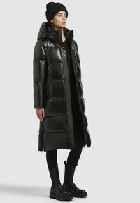 Mjuk svart pufferjacka med en glänsande yta, knälång, med huva, stora kviltade sektioner och dragkedjefickor, i kombination med svarta stövlar.