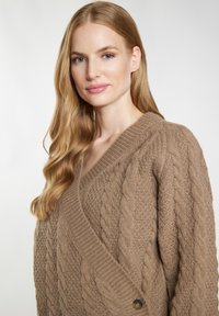 Beige kabelgebreide trui met een diepe V-hals, textuurstof en een knopelement aan de zijkant. Zachte, chunky ontwerp ideaal voor lagen.