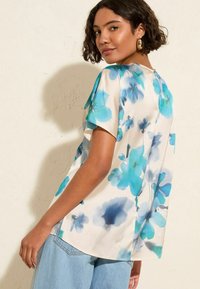 Blusa floral en tejido suave con flores azules y turquesas sobre un fondo crema. Mangas cortas y un corte holgado con aberturas laterales.
