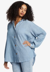 Light blue long-sleeve button-up shirt with vertical white stripes, two front pockets, and rounded hemline. Fabric appears soft and textured.