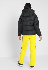 Chaqueta de plumas negra con capucha, con un diseño acolchado y puños elásticos. Combinada con pantalones de chándal amarillos brillantes y zapatillas negras.