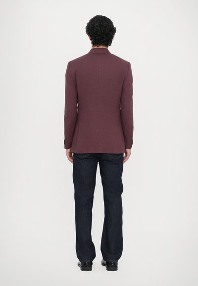 Burgunderfarbener Blazer mit taillierter Passform, einfacher Schlitze und gemustertem Stoff, kombiniert mit dunkelblauen Jeans und schwarzen Schuhen, von hinten gezeigt.