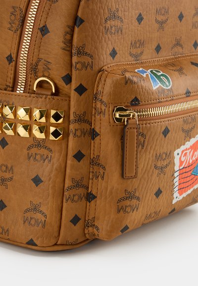 Sac en cuir brun texturé arborant un motif de logo MCM noir, des clous pyramide dorés et une poche zippée avec des accents de patch colorés.