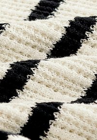 Krémová pletená textília s textúrovaným čiernym zigzag vzorom v detailnom pohľade.