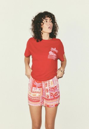 Žena s kučeravými vlasmi, oblečená v červenom tričku s potlačou palmy a farebných vzorovaných šortkách, stojaca pred jednoduchým pozadím.