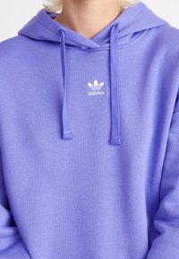 Světle fialová mikina s klokankou a šňůrkami na stažení. Na hrudi se nachází malé bílé logo Adidas. Měkká textilie.