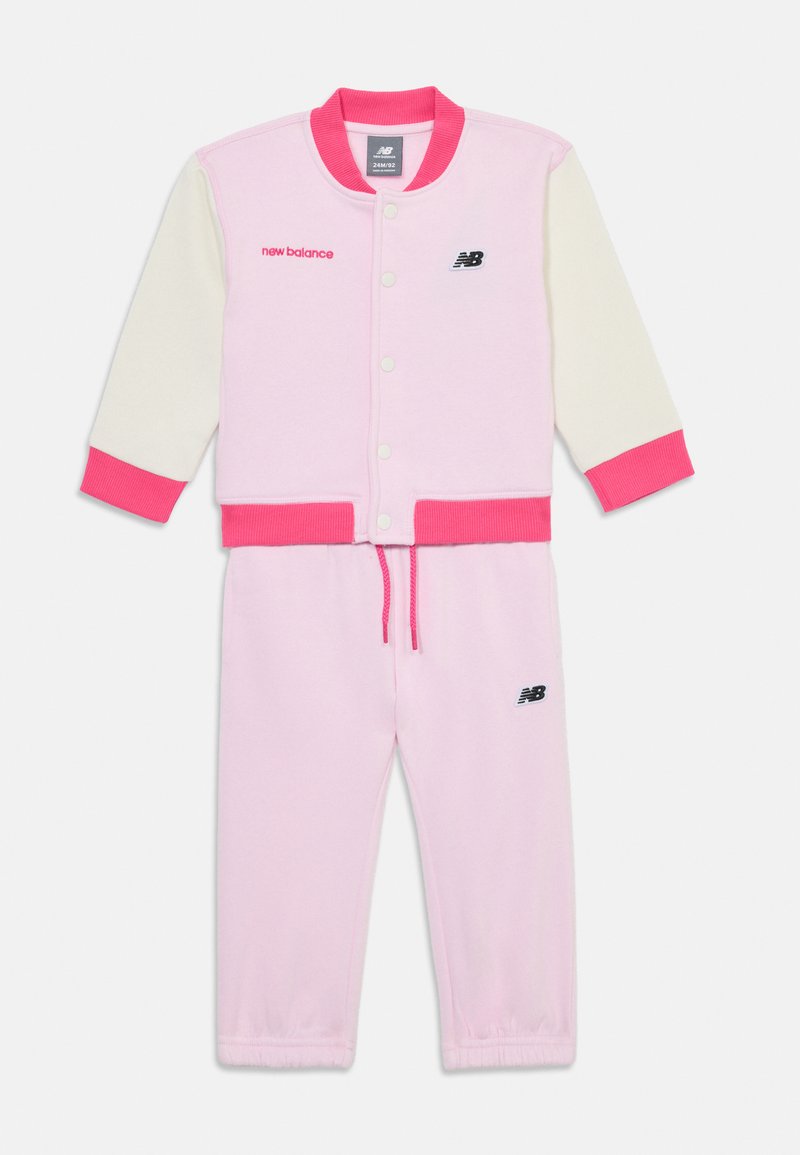 Ensemble de sport rose et crème comprenant une veste à boutons avec des poignets contrastants et un pantalon de survêtement assorti. Logos brodés en noir.
