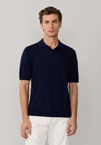 Joven con cabello castaño claro que lleva una camisa tipo polo de manga corta azul marino y pantalones blancos, posando frente a un fondo claro y sencillo.