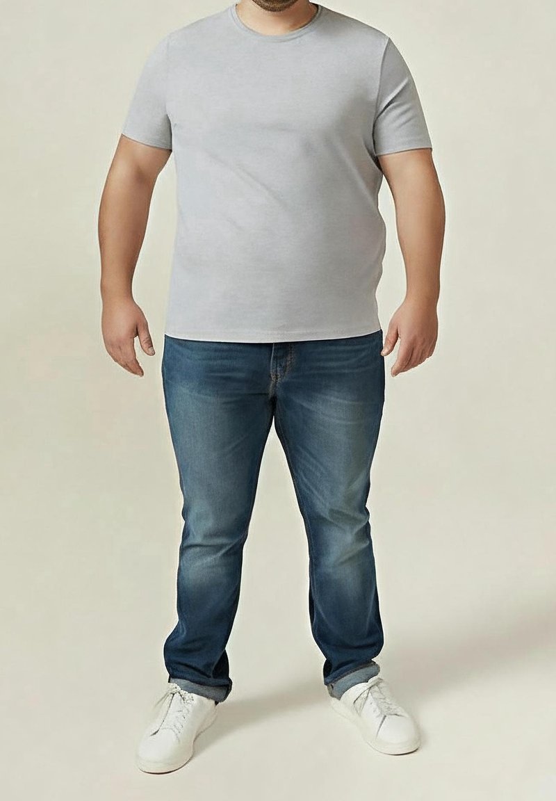 Homme debout portant un t-shirt manches courtes gris clair, un jean bleu et des baskets blanches sur un fond uni.