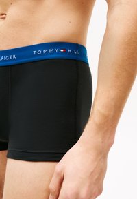 Boxer shorts negros con una cinturilla elástica azul que presenta el logo "TOMMY HILFIGER". Textura suave, diseño ajustado y longitud a mitad del muslo.