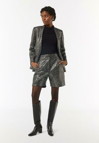 Lśniąca czarna i srebrna marynarka z dopasowanymi szortami, w zestawieniu z czarnym golfem i czarnymi skórzanymi kozakami do kolan.