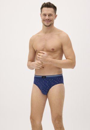 Hombre sonriente de pie, vistiendo calzoncillos azules con estampado, con las manos entrelazadas cerca del pecho, sobre un fondo claro y liso.