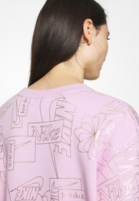 Rosa sweatshirt med guldfolie "Nike"-logotyper och mönster. Mjuk tyg med rundad halsringning. Synlig stor hoopsörhänge.