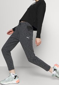 Osoba przechylająca się do tyłu, ubrana w czarną krótką bluzę z kapturem, szare dresowe spodnie Puma, białe skarpetki oraz jasnoszare sneakersy z brzoskwiniowymi akcentami.