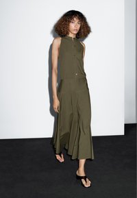 Olijfgroene, mouwloze jurk met een knoopsluiting aan de voorkant en een gelaagde rok. Glad materiaal, vloeiende silhouet met een subtiele A-lijn vorm.